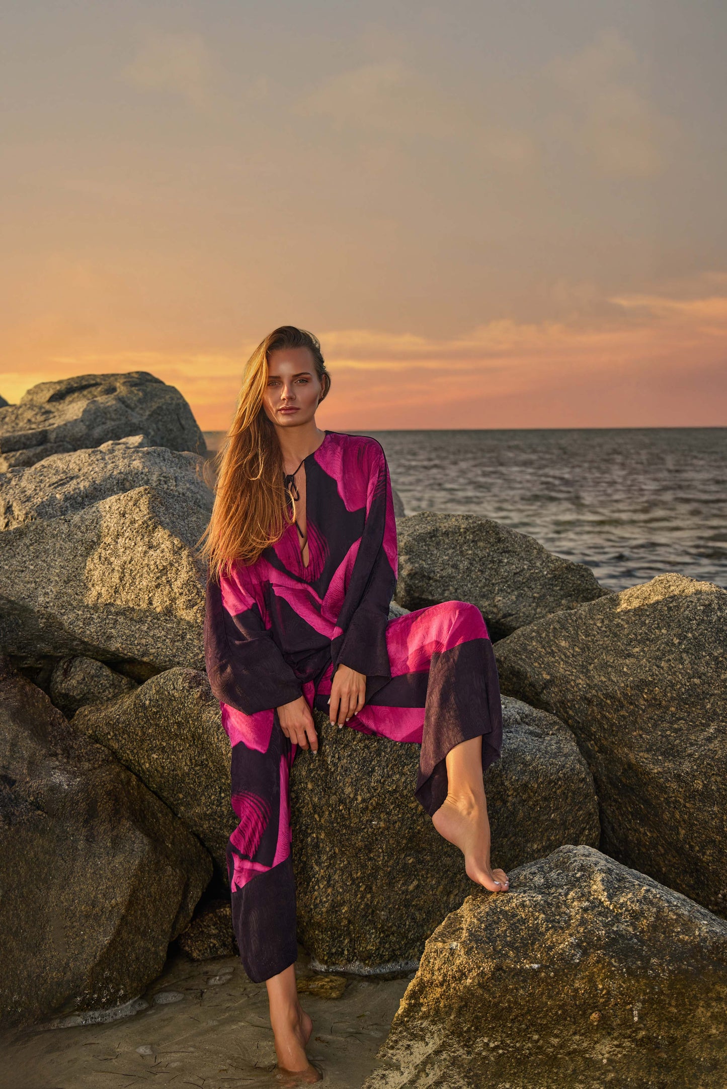 Woman in a pink and black outfit sitting on rocks by the ocean at sunset.