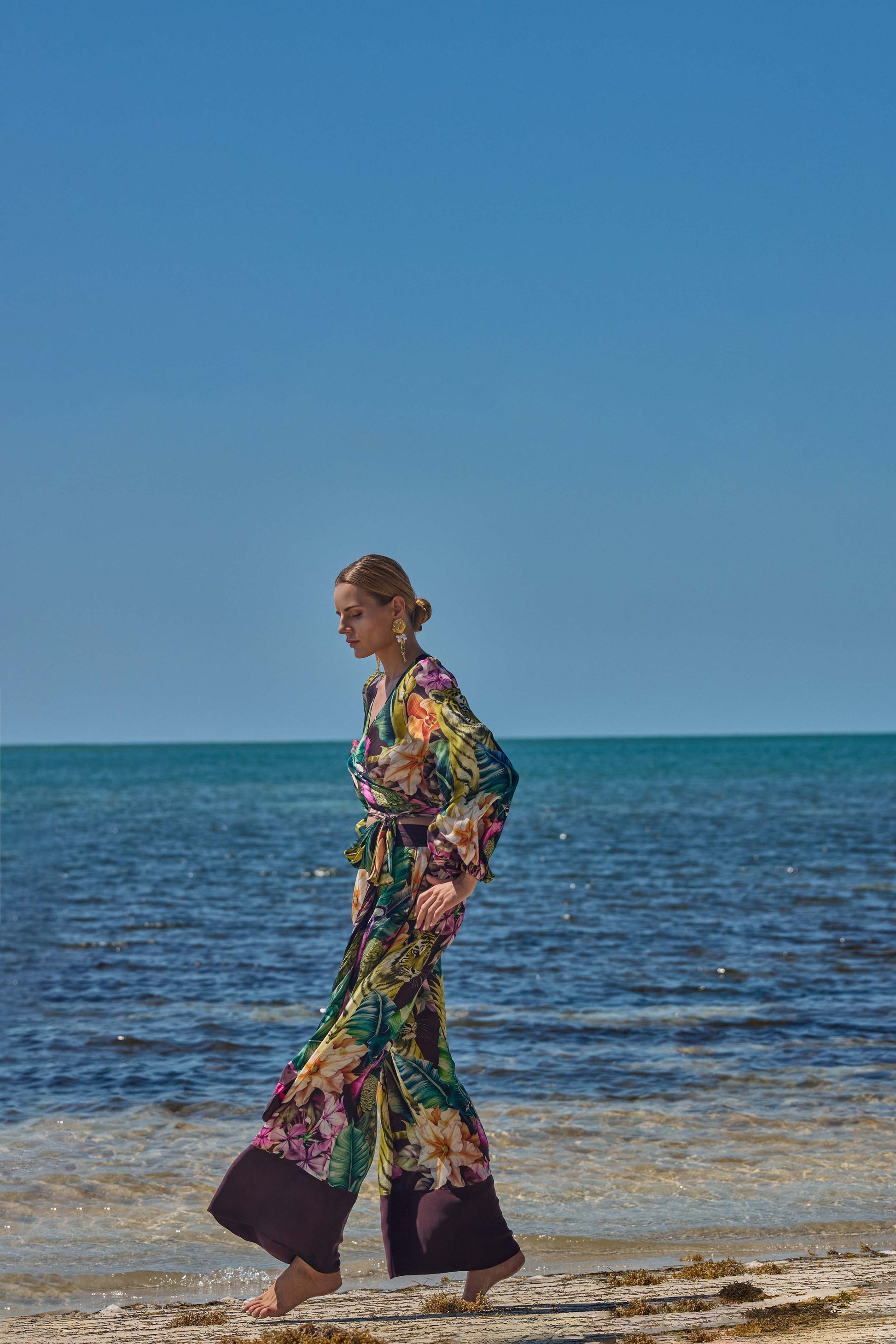Woman in a colorful floral outfit walking on a beach with clear blue sky and ocean.