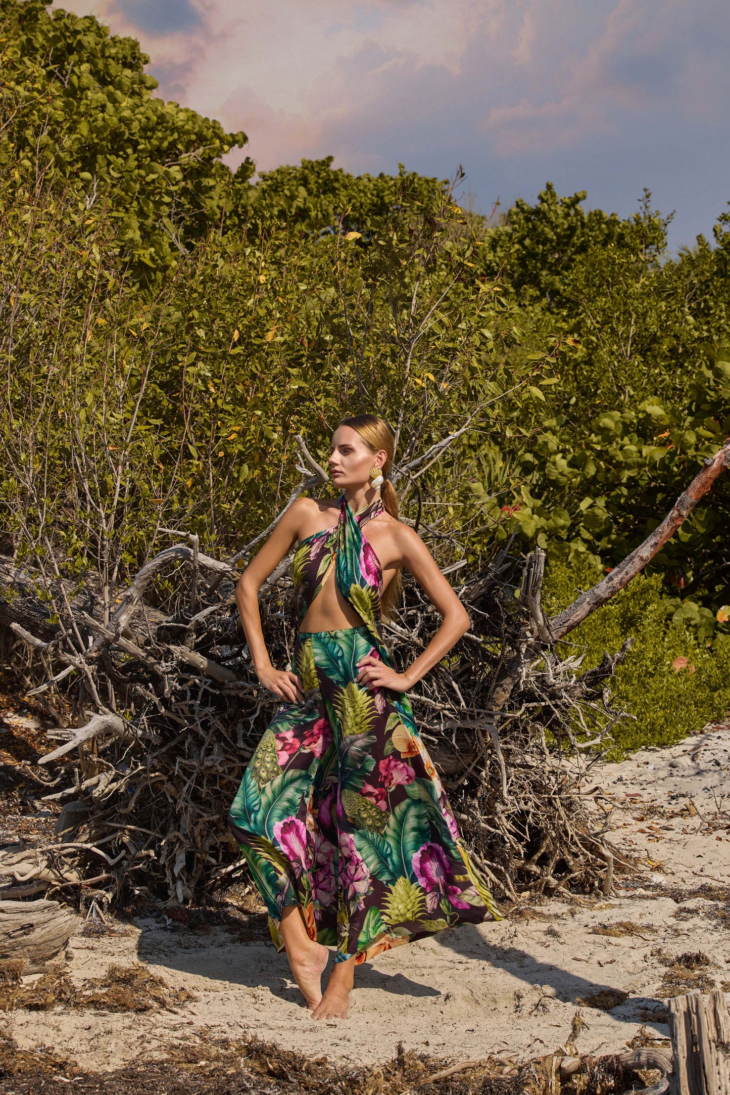 Woman in a floral outfit standing on a sandy beach with greenery in the background