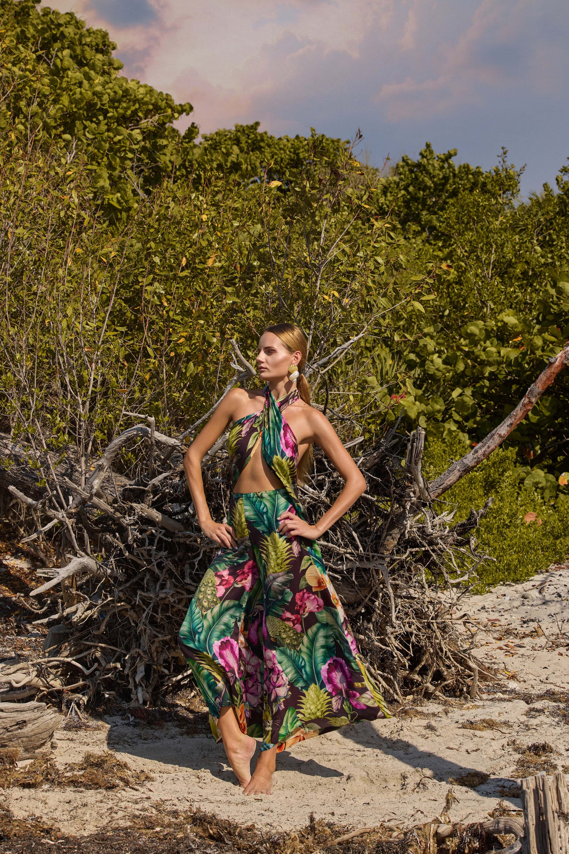 Woman in a floral outfit standing on a sandy beach with greenery in the background
