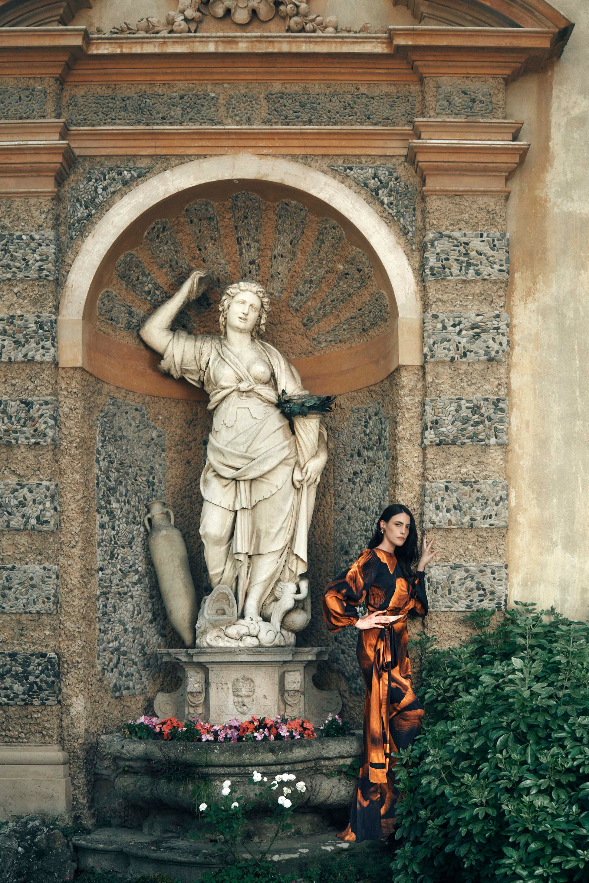 Woman in an orange and black outfit standing next to a classical statue in an outdoor setting.